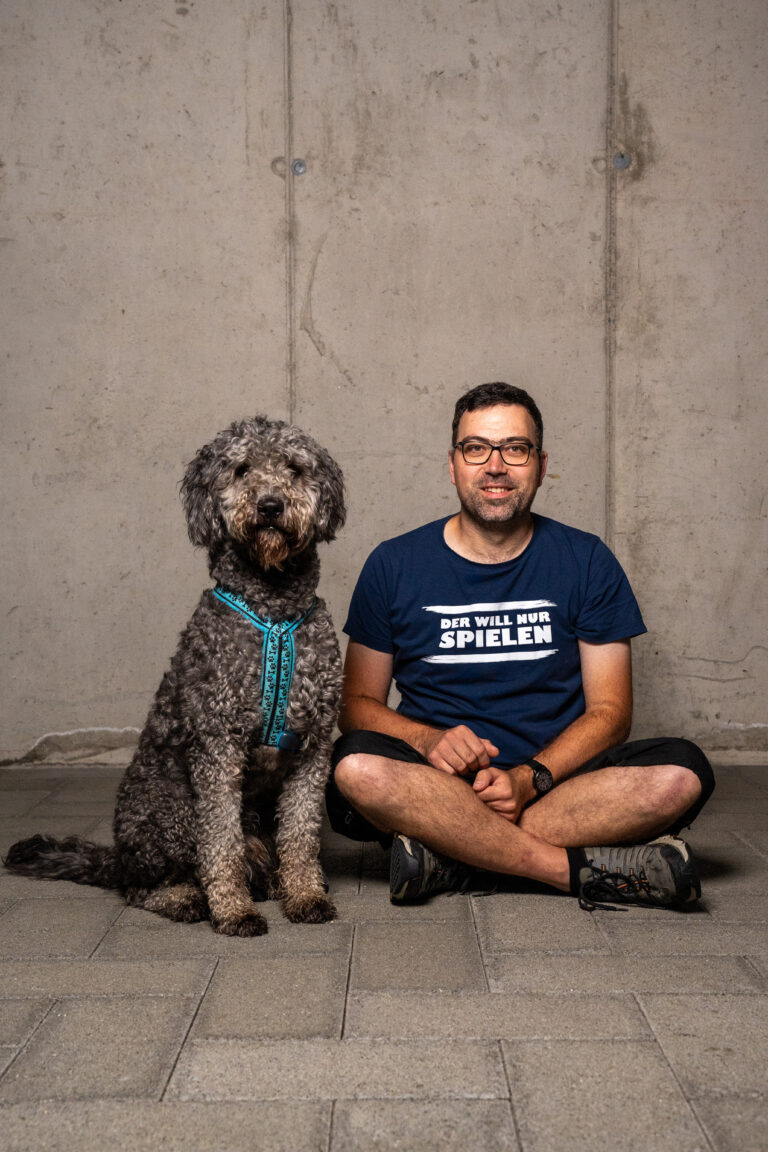 Mann mit Brille und blauem Tshirt mit weissem Spruch darauf sitzt im Schneidersitz am Boden rechts neben ihm sitzt ein grauer, größer Hund mit lockigem Fell beide schauen direkt in die Kamera