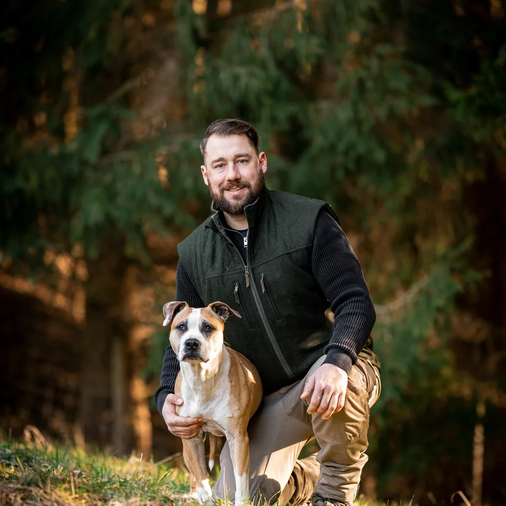 DOGS Hundetrainer Andreas Gomsi kniet neben stehendem Staffordshireterrier mit einem Bein am Boden und legt seinen rechten Arm um den Hund